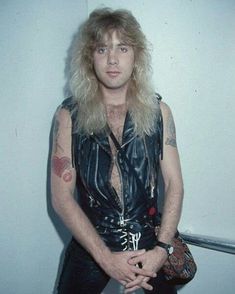 a man with long blonde hair and leather vest posing for a photo in front of a white wall