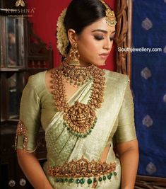 a woman in a green and gold saree with jewelry on her neck, standing next to a red wall