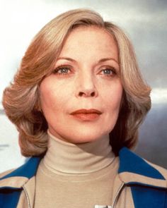 an older woman with blonde hair wearing a blue jacket and tan turtle neck sweater is looking at the camera