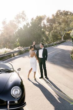 a bride and groom walking down the street in front of a black car with their arms around each other