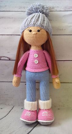 a crocheted doll with red hair wearing blue pants and a pink shirt is standing on a white wooden floor