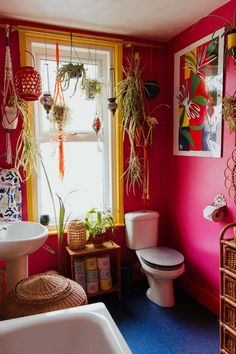 a bathroom with pink walls and plants hanging from the window above the toilet, sink and bathtub