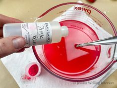 a person is holding a tube of red liquid in front of a mixing bowl filled with pink dye