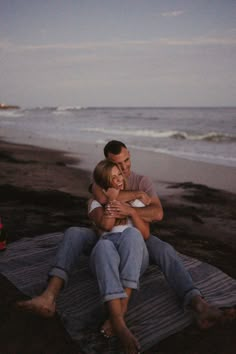 a man and woman are sitting on a blanket at the beach, hugging each other