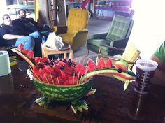 a watermelon bowl filled with fruit on top of a wooden table next to two people