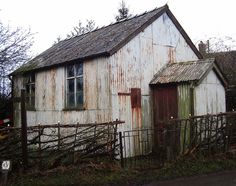 an old white barn with rusted paint on it's roof and door is next to a fence