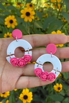 a hand holding two white and pink earrings with flowers on them, in front of yellow flowers