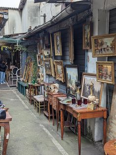 there are many paintings on the wall and tables in front of this building that is covered with framed pictures