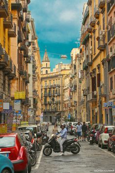 people are riding motorcycles in the middle of an old city street lined with parked cars