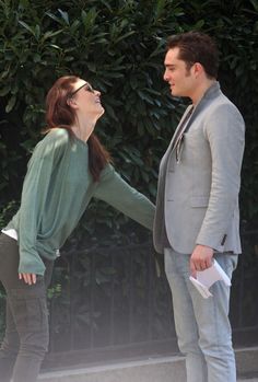 a man and woman standing next to each other in front of a fence with trees