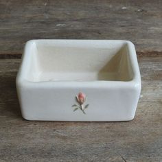 a small white square bowl with a rose on the front and bottom, sitting on a wooden surface