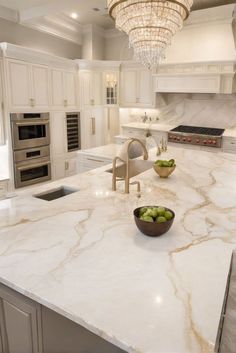 a large kitchen with marble counter tops and white cabinets, an island in the middle has two bowls of fruit on it