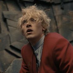 a man with blonde hair wearing a red jacket and tie in front of a rock wall