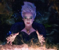 a woman with purple hair and green eyes is surrounded by fire