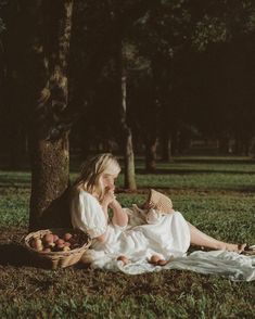 a woman sitting in the grass holding a baby next to a basket full of apples