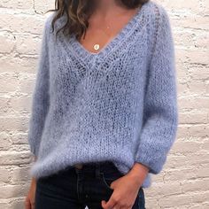 a woman standing in front of a white brick wall wearing a blue sweater and jeans