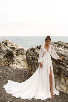 a woman standing on top of a rocky beach next to the ocean wearing a long white dress