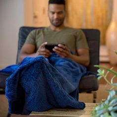 a man sitting in a chair holding a cell phone and looking at it with a blue blanket on his lap