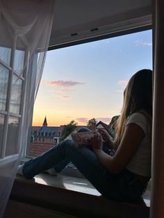 a woman sitting on a window sill reading a book in the sun set behind her