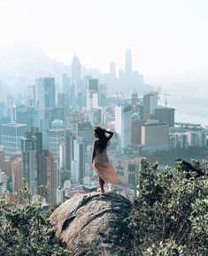 a woman standing on top of a large rock in front of a cityscape