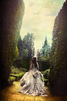 a woman in a white dress is walking through some bushes and trees with a castle in the background