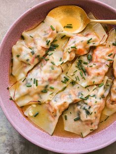 a pink bowl filled with ravioli covered in sauce and topped with parsley next to a spoon