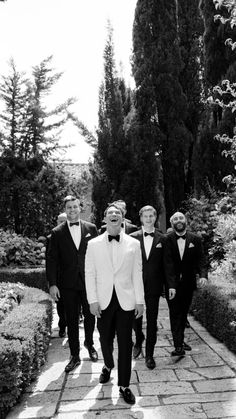 a group of men in tuxedos walking down a brick path with trees and bushes behind them