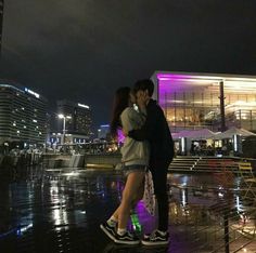 a man and woman kissing in front of a building on a rainy night with lights reflecting off the water