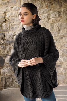 a woman standing in front of a stone wall wearing a black cable knit sweater and jeans