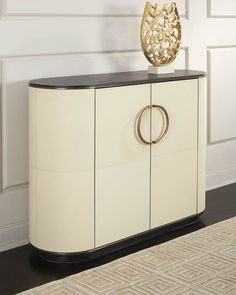 a white and black cabinet with a gold vase on it's top, in front of a wall