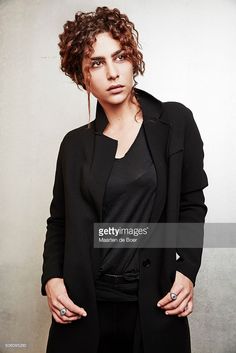 a woman with curly hair wearing a black jacket posing for a photo in front of a white wall