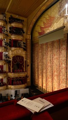 a red couch sitting in front of a window with curtains on the wall and an open book