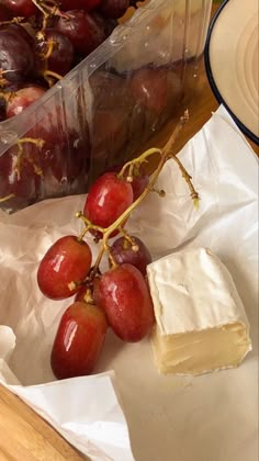 grapes and cheese are sitting on wax paper next to a plastic container filled with fruit