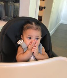 a baby sitting in a highchair holding something up to her mouth and looking at the camera