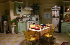 a kitchen with green cabinets and yellow chairs in front of a table that has food on it