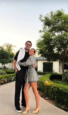 a man standing next to a woman on top of a sidewalk in front of trees