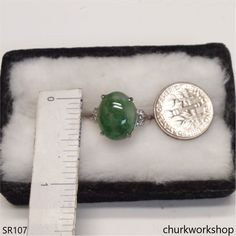 a green jade and diamond ring sitting on top of a white velvet box next to a ruler