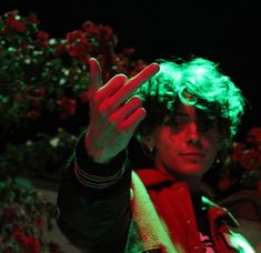 a man with green hair making the vulcan sign while standing in front of red flowers