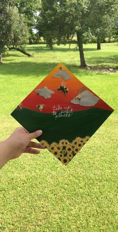 a hand holding up a colorful kite in the air with trees and grass behind it