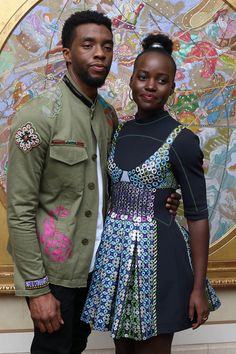 a man and woman standing next to each other in front of a wall with paintings on it