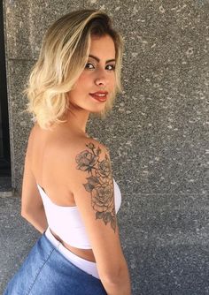 a woman with a tattoo on her arm standing in front of a wall and looking at the camera