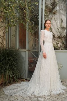 a woman standing in front of a window wearing a wedding dress with long sleeves and beading