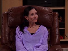 a woman sitting in a brown recliner wearing a purple shirt and pearls on her necklace