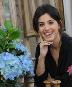 a woman sitting at a table with flowers in front of her and smiling for the camera