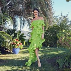 a woman in a green dress standing on grass next to palm trees and potted plants