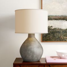 a table lamp sitting on top of a wooden dresser next to a painting and bowl