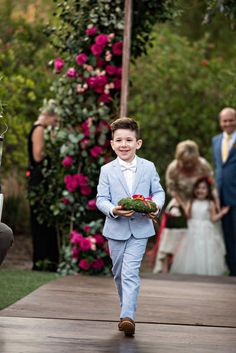 a young boy in a blue suit walking down the aisle at a wedding with other people