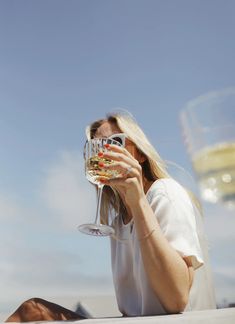 a woman sitting on the ground holding a wine glass up to her face and looking into the distance