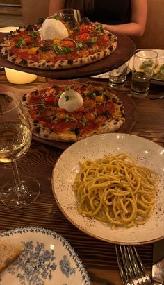 two pizzas and some pasta are on the table with glasses of wine in front of them