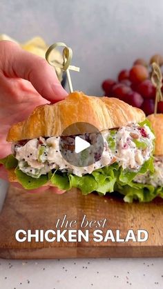 a person is holding a chicken salad sandwich on a cutting board with grapes in the background